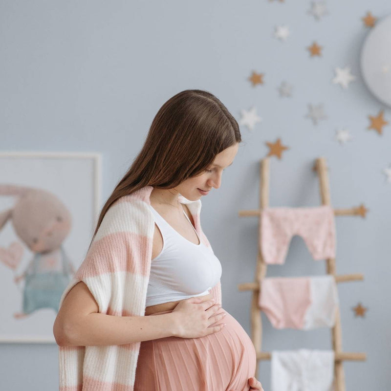 A pregnant woman gently holds her baby bump with one hand while draping a striped shawl over her shoulders. She stands in a softly lit room decorated with star-shaped wall hangings, a ladder holding baby clothes, and a framed picture of a bunny.