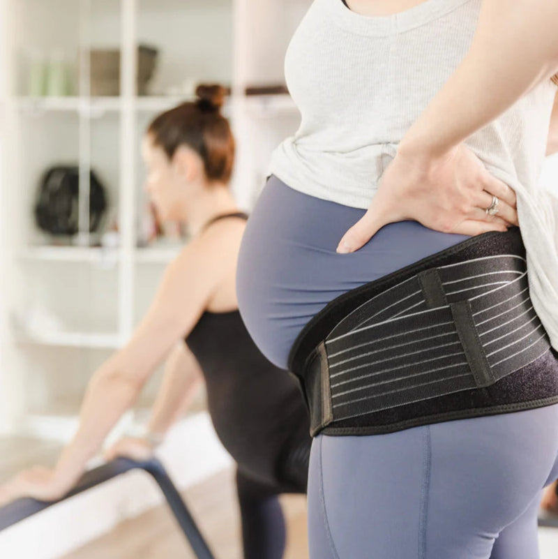 A pregnant person wearing the GRDL Pregnancy Belt β Trusted Pelvic & Back Support for Pregnancy by GRDL and workout clothes stands in the foreground, while another person exercises in a bright, home-like background.