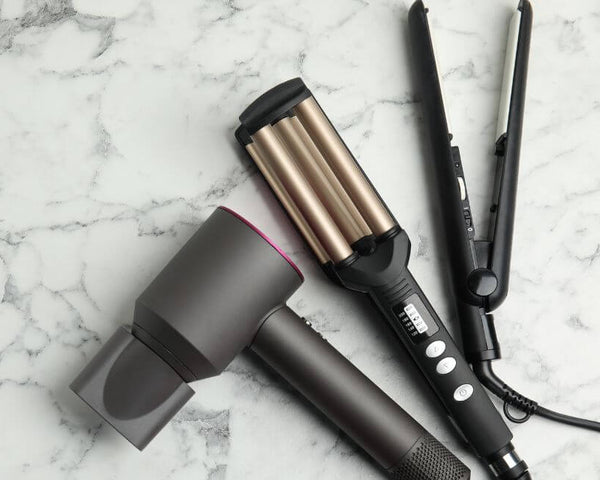 A hair styling set on a marble surface, featuring a gray hairdryer, a black and gold hair waver, and a black hair straightener.