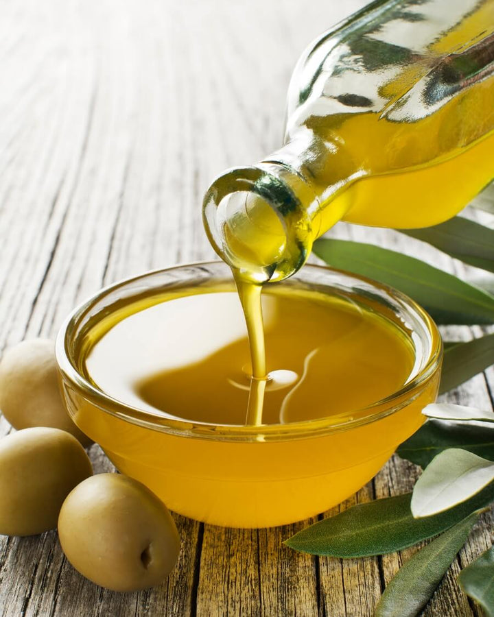 A glass bottle is pouring olive oil into a small glass bowl on a wooden surface. Two whole olives and olive leaves are placed beside the bowl, enhancing the fresh, natural aesthetic.