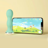 A teal, wavy Perifit Kegel Exerciser – Smart Pelvic Floor Trainer stands upright beside a smartphone showing an animated landscape game, set against a light yellow background for pelvic floor strengthening.