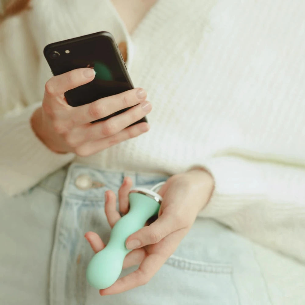 A person in a white sweater and blue jeans holds a smartphone in one hand and a mint green Perifit Kegel Exerciser – Smart Pelvic Floor Trainer by Perifit in the other.