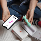 A person sits cross-legged on a bed, holding the mint green Perifit Kegel Exerciser – Smart Pelvic Floor Trainer in one hand and a smartphone showing the same product in the other, with the Perifit box and lid nearby.