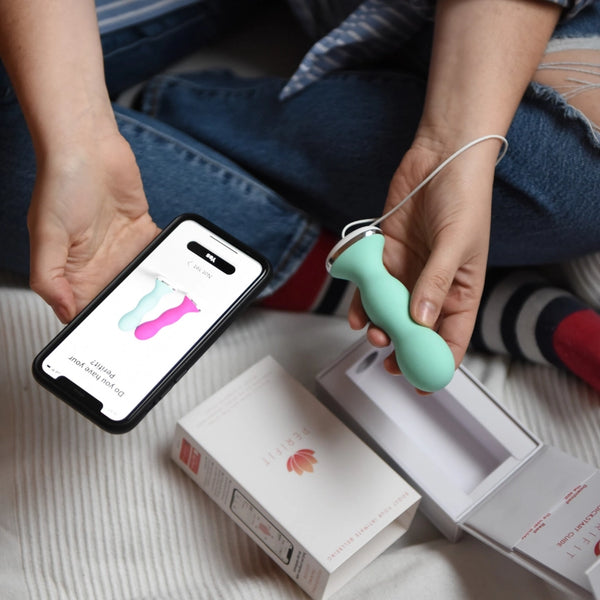 A person sits cross-legged on a bed, holding the mint green Perifit Kegel Exerciser – Smart Pelvic Floor Trainer in one hand and a smartphone showing the same product in the other, with the Perifit box and lid nearby.