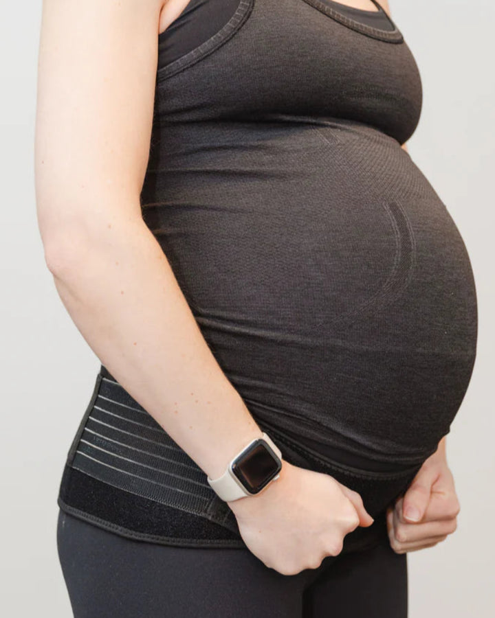 A pregnant person wearing a dark tank top, pants, and a smartwatch stands sideways, holding the GRDL Pregnancy Belt for trusted pelvic and back support by GRDL near their waist to support their baby bump.