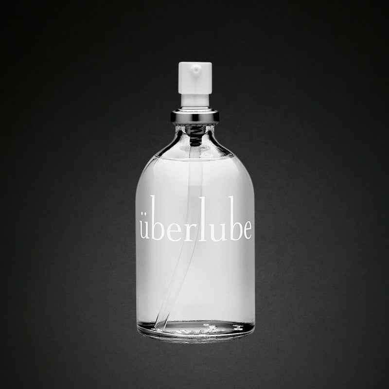 A clear glass bottle labeled "überlube" with a silver neck and a white pump dispenser, set against a dark background.