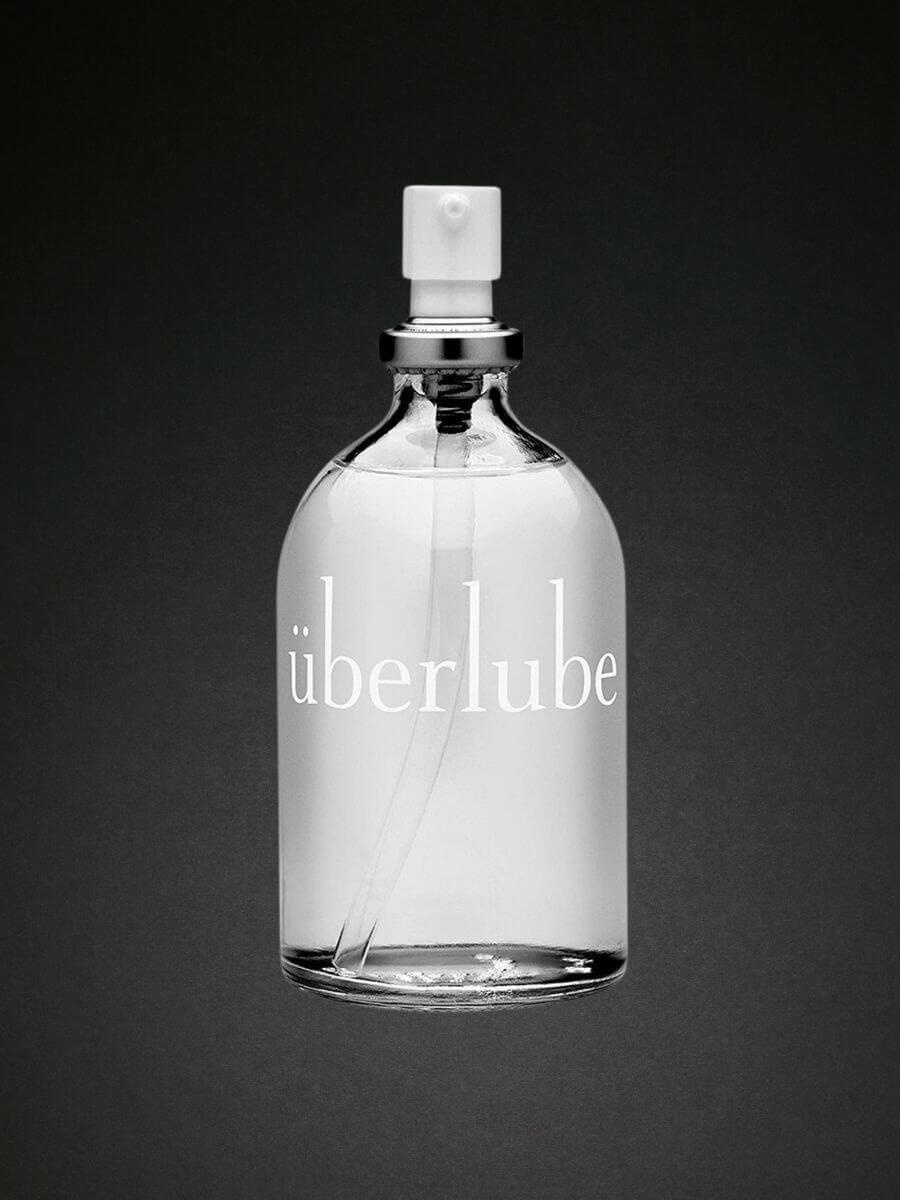 A clear glass bottle with the word "überlube" written on it, featuring a silver and white spray dispenser. The background is a gradient of dark to light gray.