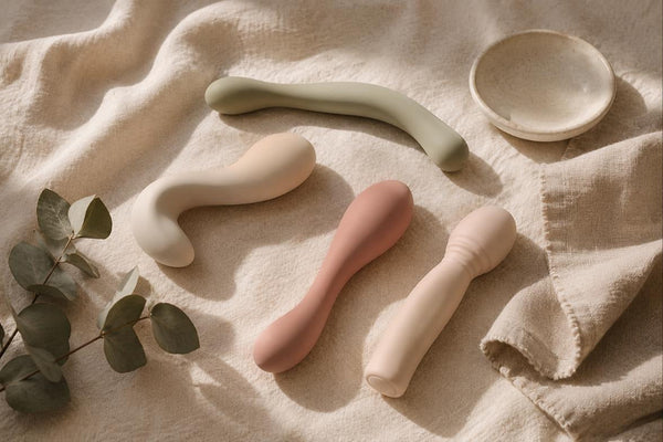 Flat-lay of four smooth silicone vibrators in blush, cream and sage tones on soft linen with a eucalyptus sprig