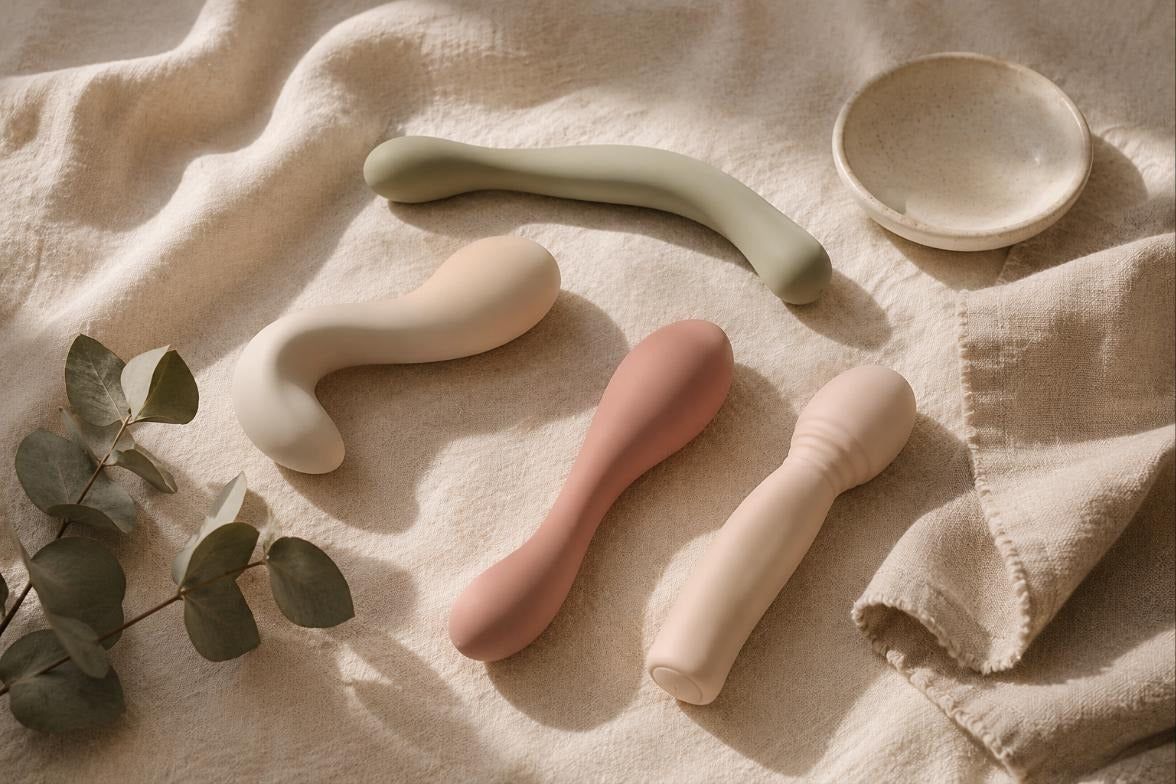 Flat-lay of four smooth silicone vibrators in blush, cream and sage tones on soft linen with a eucalyptus sprig