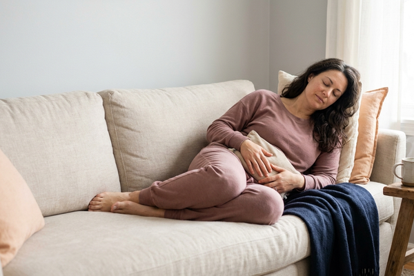 Australian woman resting on a sofa at home holding a warm heat pack against her lower abdomen to ease pelvic pain