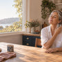 Woman around 50 sitting at a sunlit kitchen table at home, eyes closed, holding a glass of cold water against her neck