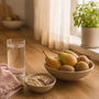 Glass of water, bowl of pears and kiwifruit and a bowl of oats with linseed on a sunlit kitchen bench