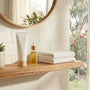 Bathroom shelf with a plain cosmetic tube, an amber dropper bottle, folded towels and a succulent below a round mirror