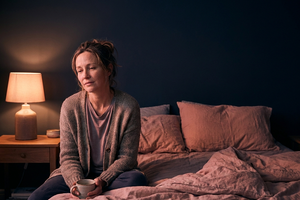 Woman sitting on the edge of her bed by a warm lamp at night holding a mug, awake with nocturia