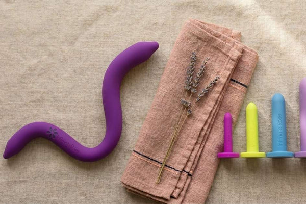 Intimate Rose pelvic wand beside a graduated set of coloured vaginal dilators on linen, comparing the two pelvic floor tools