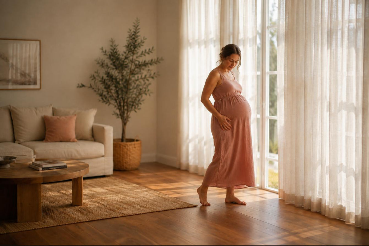 Pregnant woman standing by a sunlit window at home with a hand low on the side of her bump, easing round ligament pain