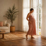 Pregnant woman standing by a sunlit window at home with a hand low on the side of her bump, easing round ligament pain