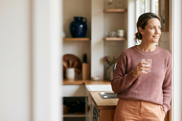 Woman holding a glass of water by a sunlit kitchen window at home, calm — urge incontinence versus stress incontinence