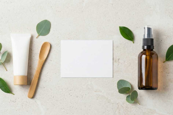 Flat-lay on a pale stone surface of a white cosmetic tube with a bamboo applicator and an amber pump bottle among eucalyptus leaves, representing a vaginal moisturiser and a lubricant side by side