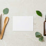 Flat-lay on a pale stone surface of a white cosmetic tube with a bamboo applicator and an amber pump bottle among eucalyptus leaves, representing a vaginal moisturiser and a lubricant side by side