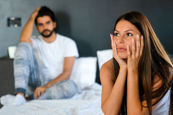 Woman sitting on the edge of a bed looking anxious with her partner behind her, reflecting the strain vaginismus can place on intimacy