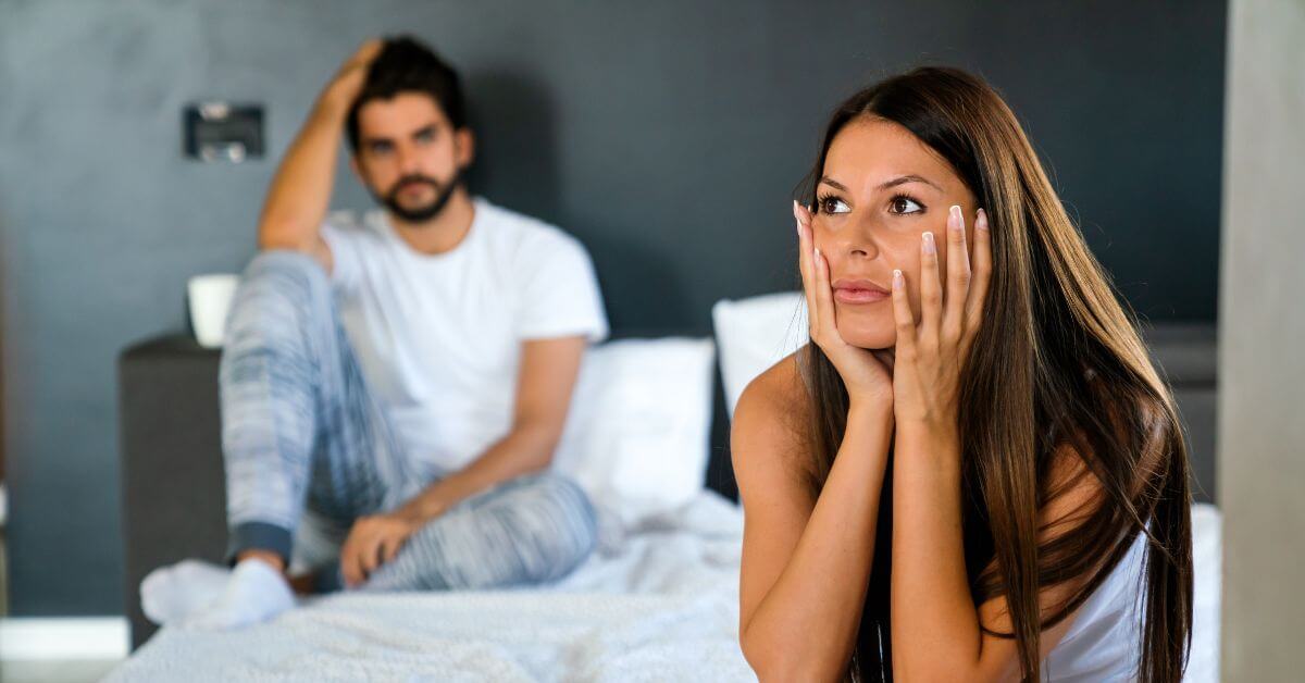 Woman sitting on the edge of a bed looking anxious with her partner behind her, reflecting the strain vaginismus can place on intimacy
