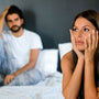 Woman sitting on the edge of a bed looking anxious with her partner behind her, reflecting the strain vaginismus can place on intimacy