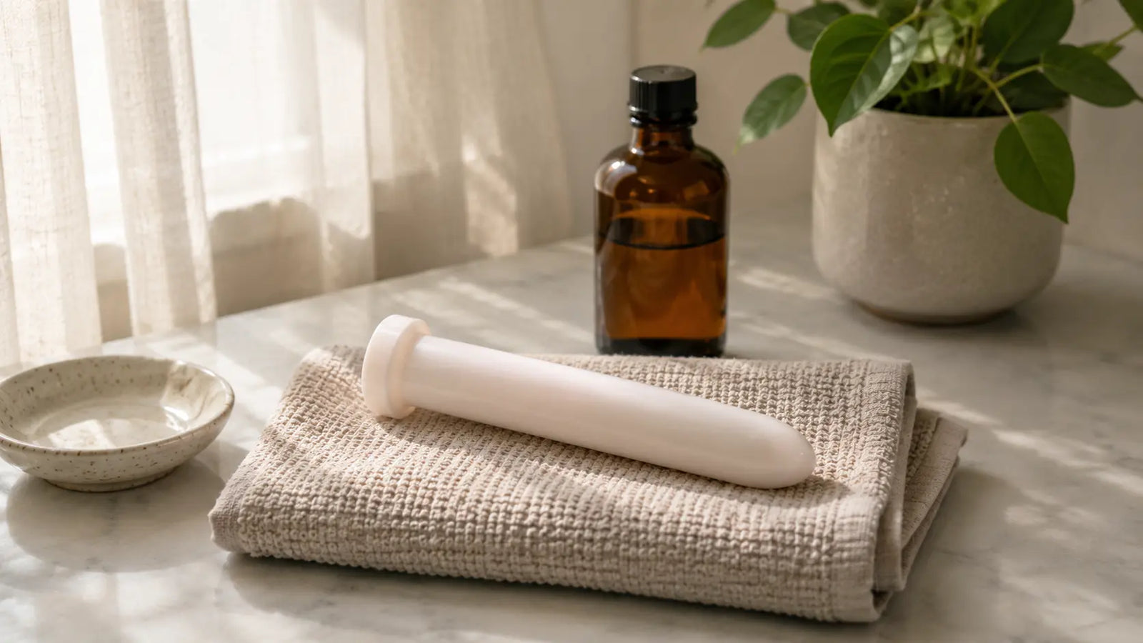White plastic Amielle Care vaginal dilator resting on a folded washcloth on marble, beside an amber-glass bottle and leafy plant — calm setting for introducing dilator therapy.