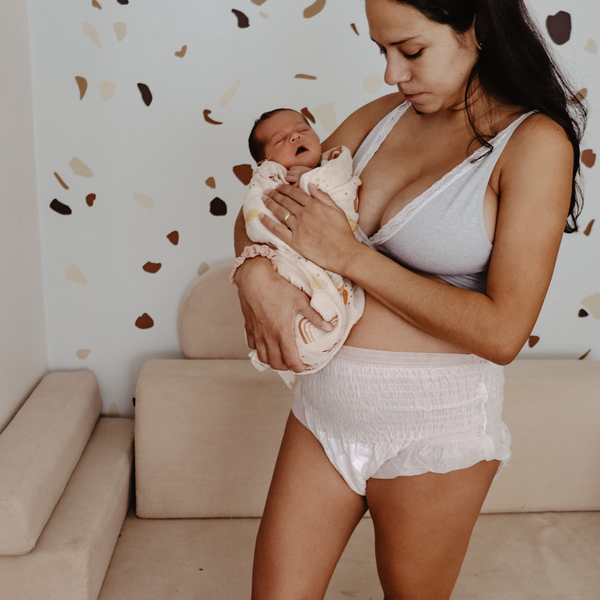 A woman in a gray tank top and Bubba Bump Disposable Postpartum Underwear cradles a swaddled newborn in a cozy room with a patterned wall, gazing tenderly at the baby by a beige sofa, embracing moments of C-section recovery.