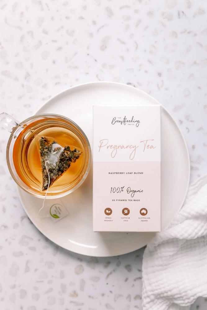 A cup of Raspberry Leaf Tea with a pyramid tea bag inside sits on a white saucer next to a box labeled "The Breastfeeding Tea Co - Pregnancy Tea - Raspberry Leaf Blend - 100% Organic." The box features icons indicating organic ingredients, caffeine-free, and eco-friendly packaging, perfect with pregnancy-safe herbs.