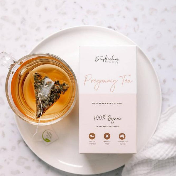 A cup of Raspberry Leaf Tea with a pyramid tea bag inside sits on a white saucer next to a box labeled "The Breastfeeding Tea Co - Pregnancy Tea - Raspberry Leaf Blend - 100% Organic." The box features icons indicating organic ingredients, caffeine-free, and eco-friendly packaging, perfect with pregnancy-safe herbs.