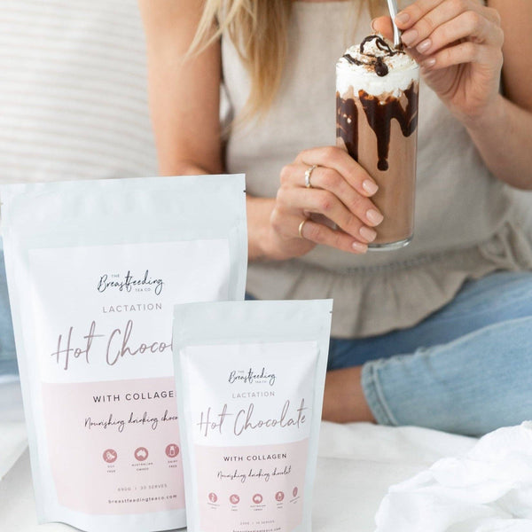 A woman is holding a glass of iced chocolate drink topped with whipped cream and chocolate drizzle. In front of her, on a white surface, are two bags of The Breastfeeding Tea Co's Lactation Hot Chocolate with Collagen, designed to support breastmilk supply—one large and one small.