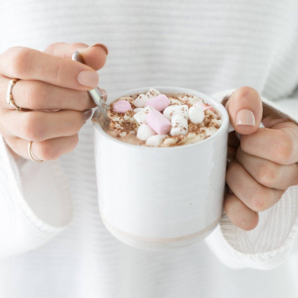 A person holding a white mug filled with Lactation Hot Chocolate with Collagen from The Breastfeeding Tea Co, topped with small pink and white marshmallows and chocolate shavings, stirring it with a spoon. The person is wearing a cozy white sweater and has neatly manicured nails.