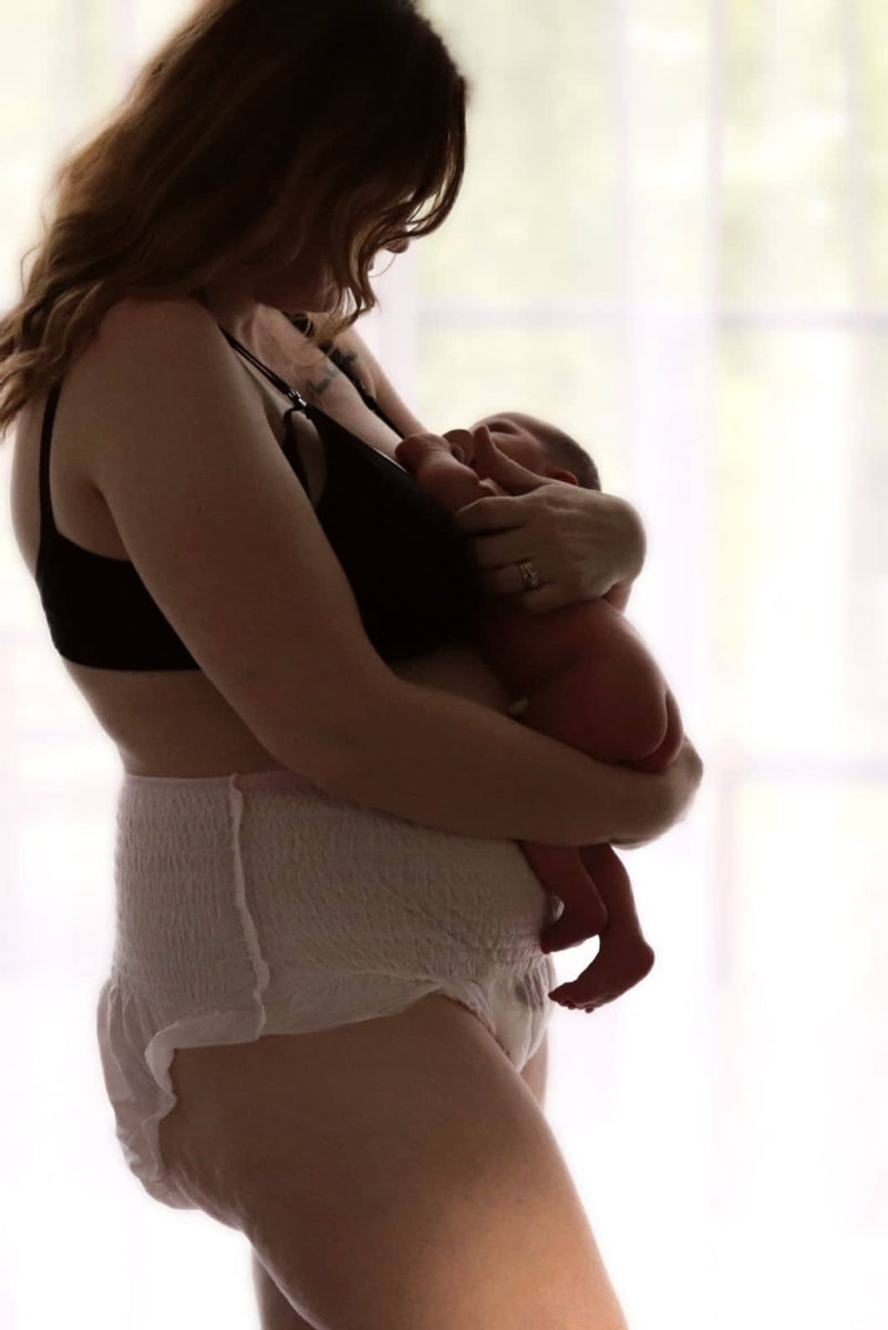 A woman with long hair stands in profile, holding her newborn close. Dressed in Bubba Bump's black bra and Disposable Postpartum Underwear, she enjoys soft, natural light from the window, creating a serene atmosphere with leak-proof protection at its finest.