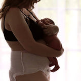 A woman with long hair stands in profile, holding her newborn close. Dressed in Bubba Bump's black bra and Disposable Postpartum Underwear, she enjoys soft, natural light from the window, creating a serene atmosphere with leak-proof protection at its finest.