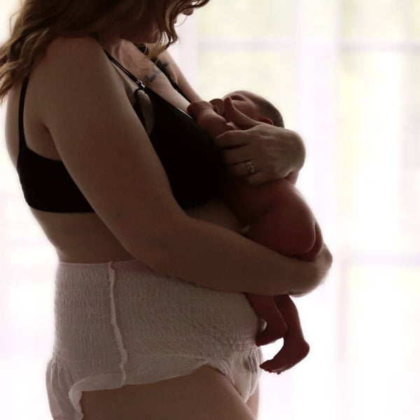 A woman with long hair stands in profile, holding her newborn close. Dressed in Bubba Bump's black bra and Disposable Postpartum Underwear, she enjoys soft, natural light from the window, creating a serene atmosphere with leak-proof protection at its finest.