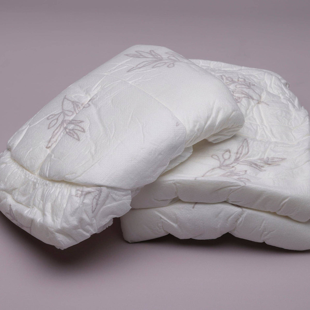 Two Bubba Bump Disposable Postpartum Underwear with floral patterns are folded neatly on a smooth, light purple surface. They feature a quilted design for softness and offer leak-proof protection to keep you comfortable and confident.