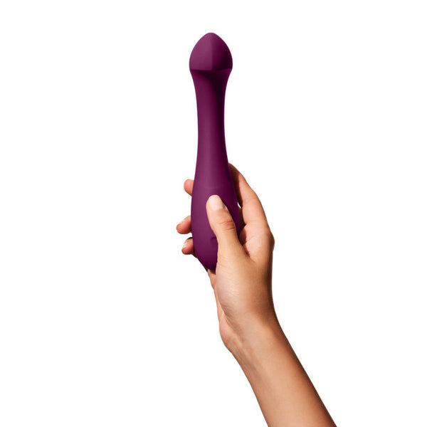 A hand holding a purple, elongated object against a white background. This object is the Dame Arc, a G-Spot vibrator made of medical-grade silicone that features a smooth, tapered shape, likely designed for clitoral stimulation.