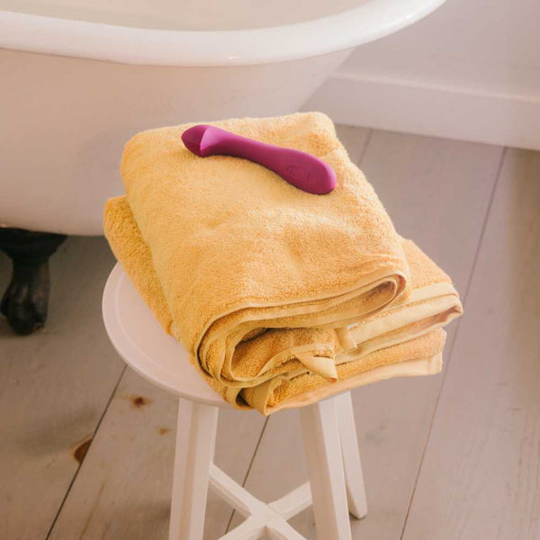 A folded yellow towel with a purple Dame Arc G-Spot Vibrator, crafted from medical-grade silicone, rests on a white stool next to the bathtub in the bathroom.