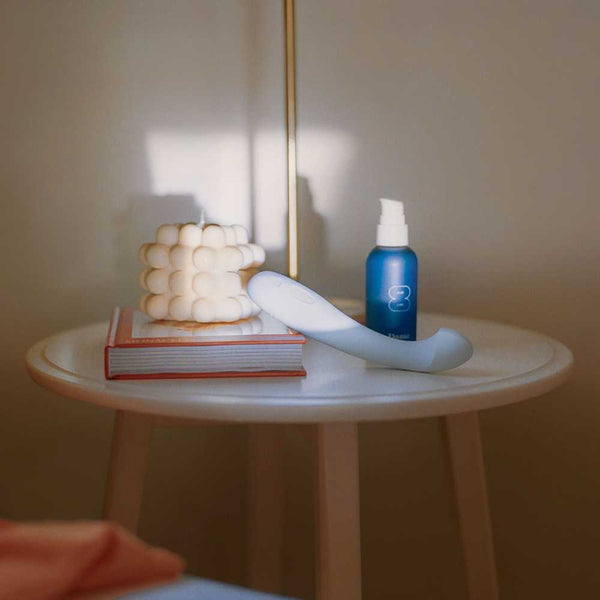 A round white side table features a decorative cube-shaped candle, a book, the Dame Arc - G-Spot Vibrator crafted from medical-grade silicone in white, and a blue spray bottle. Soft lighting casts shadows, creating a cozy atmosphere.