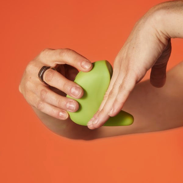 Hands holding the Kiwi Vibrating Pelvic Massager by The Pelvic People against an orange background, offering a hint of pelvic floor tightness relief.