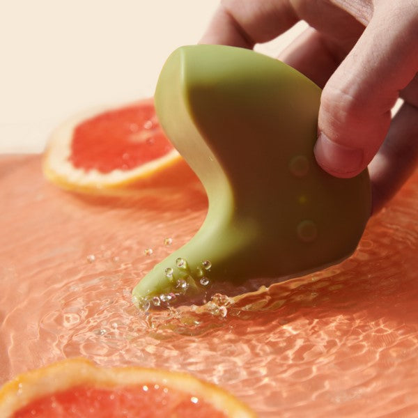 A hand holds a green, curved object above grapefruit slices with bubbles, suggesting movement in water. The Kiwi Vibrating Pelvic Massager by The Pelvic People provides pelvic tightness relief and gentle clitoral stimulation with its soothing design.
