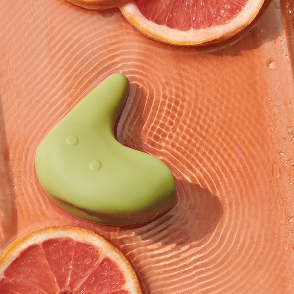 A smooth, curved green soap bar with two small indents sits in a tray of water, mirroring The Pelvic People's Kiwi Vibrating Pelvic Massager. Ripples surround grapefruit slices in the water, evoking tranquility and relief.