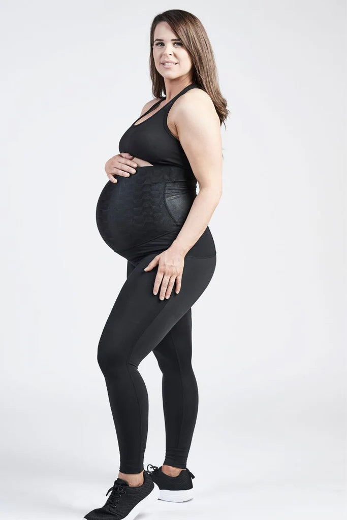 A pregnant woman stands against a plain white background, wearing a black sports bra and SRC Pregnancy Leggings - Over the Bump designed for pelvic support from SRC Health. She holds her baby bump with one hand and rests the other hand on her thigh. She has light skin, long brown hair, wears black sneakers, and is smiling.