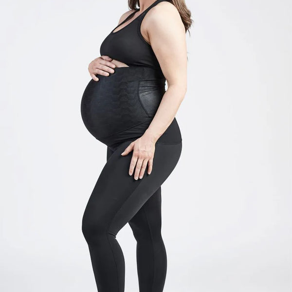 A pregnant woman stands against a plain white background, wearing a black sports bra and SRC Pregnancy Leggings - Over the Bump designed for pelvic support from SRC Health. She holds her baby bump with one hand and rests the other hand on her thigh. She has light skin, long brown hair, wears black sneakers, and is smiling.