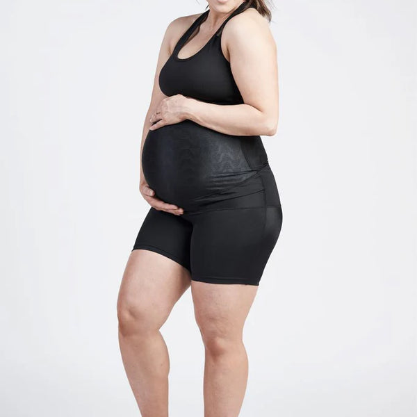 A pregnant woman in athletic wear stands with her legs slightly apart. She is wearing a black tank top and SRC Pregnancy Shorts - Mini Over the Bump by SRC Health. Her hands gently cradle her belly, and she smiles while looking down. With long, dark hair tied back in a ponytail, she seems to be embracing the moment with pelvic floor support for pain relief.