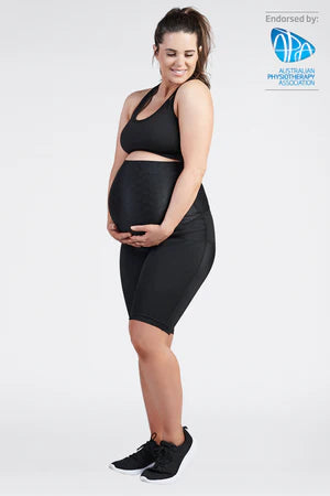 A pregnant woman, dressed in a black SRC Health sports bra and SRC Pregnancy Shorts - Over the Bump, stands with both hands gently cradling her belly. She smiles warmly as she gazes down at her bump. The image features the endorsement of the Australian Physiotherapy Association, indicated by their logo in the upper right corner.