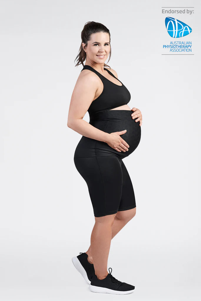 An expectant mother in a black SRC Pregnancy Shorts - Over the Bump and matching sports bra beams as she cradles her baby bump. Standing sideways in black athletic shoes, she demonstrates the pelvic support feature of the SRC Health product. The image is endorsed by the Australian Physiotherapy Association, with their logo visible in the top right corner.