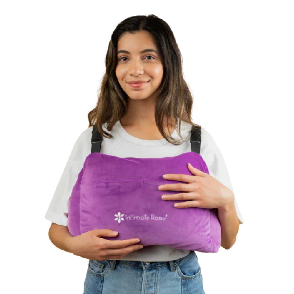 A person smiling while holding a large, purple Post-Surgery Mastectomy Pillow featuring the "Intimate Rose" logo. They are dressed in a white shirt and denim jeans.