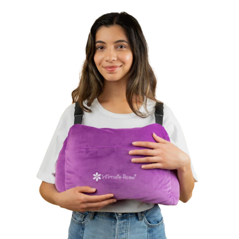 A person smiling while holding a large, purple Post-Surgery Mastectomy Pillow featuring the "Intimate Rose" logo. They are dressed in a white shirt and denim jeans.